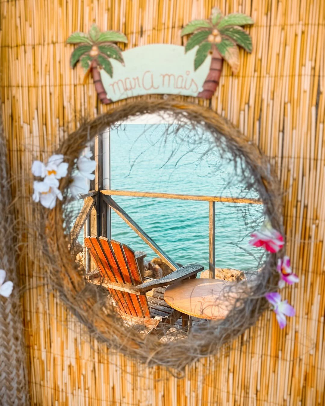 Janela redonda decorada com folhas e flores, com vista para a esplanada e o mar azul-turquesa